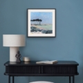 Through the Trees coastal landscape print displayed in a black frame against a blue wall above a dark console