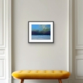Majestic Sky coastal giclée print by Louise Turnbull displayed in a black frame above a mustard yellow bench in a bright hallway.