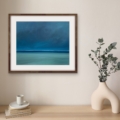 Light and Magic blue abstract seascape print displayed with a white mount and natural wood frame above a wooden console table with a ceramic vase and eucalyptus.