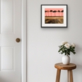 Listen Softly giclée print featuring silhouetted trees against a coral-pink sky, displayed in a slim black frame with a white mount on a white wall above a wooden stool with flowers.