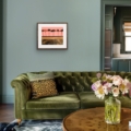 Listen Softly giclée print featuring silhouetted trees against a coral-pink sky, displayed in a wooden frame above an olive-green velvet sofa in a contemporary living room.