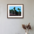 Reaching Stillness woodland giclée print featuring tall trees against a blue sky, displayed in a wooden frame with a white mount above a rustic table and vase of dried grasses.