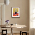 Colourful still life print with a golden green background and vibrant orange-red tabletop, shown in a wooden frame above a contemporary dining table.