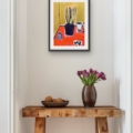 Colourful still life print with a golden green background and vivid orange-red tabletop, shown in a black frame above a rustic wooden console table.