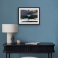 Yearning For atmospheric seascape giclée print by Louise Turnbull displayed in a black frame with white mount against a muted blue wall above a dark console table.